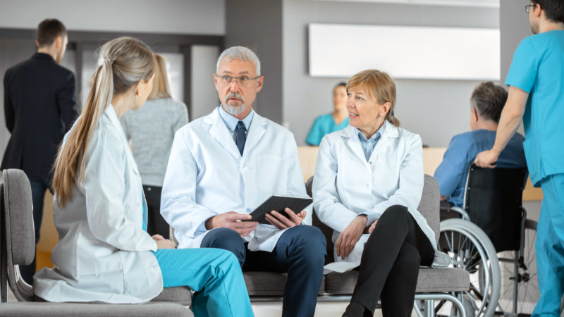 In the Lobby of the Hospital Three Professional Doctor Have Discussion while Sitting. They Use Tablet Computer. Busy Modern Hospital with Best Specialists and Medicare in Country.