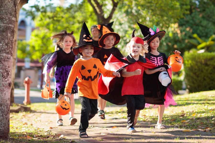 Child in Halloween costume. Mixed race Asian and Caucasian kids and parents trick or treat on street. Little boy and girl with pumpkin lantern and candy bucket. Baby in witch hat. Autumn holiday fun.